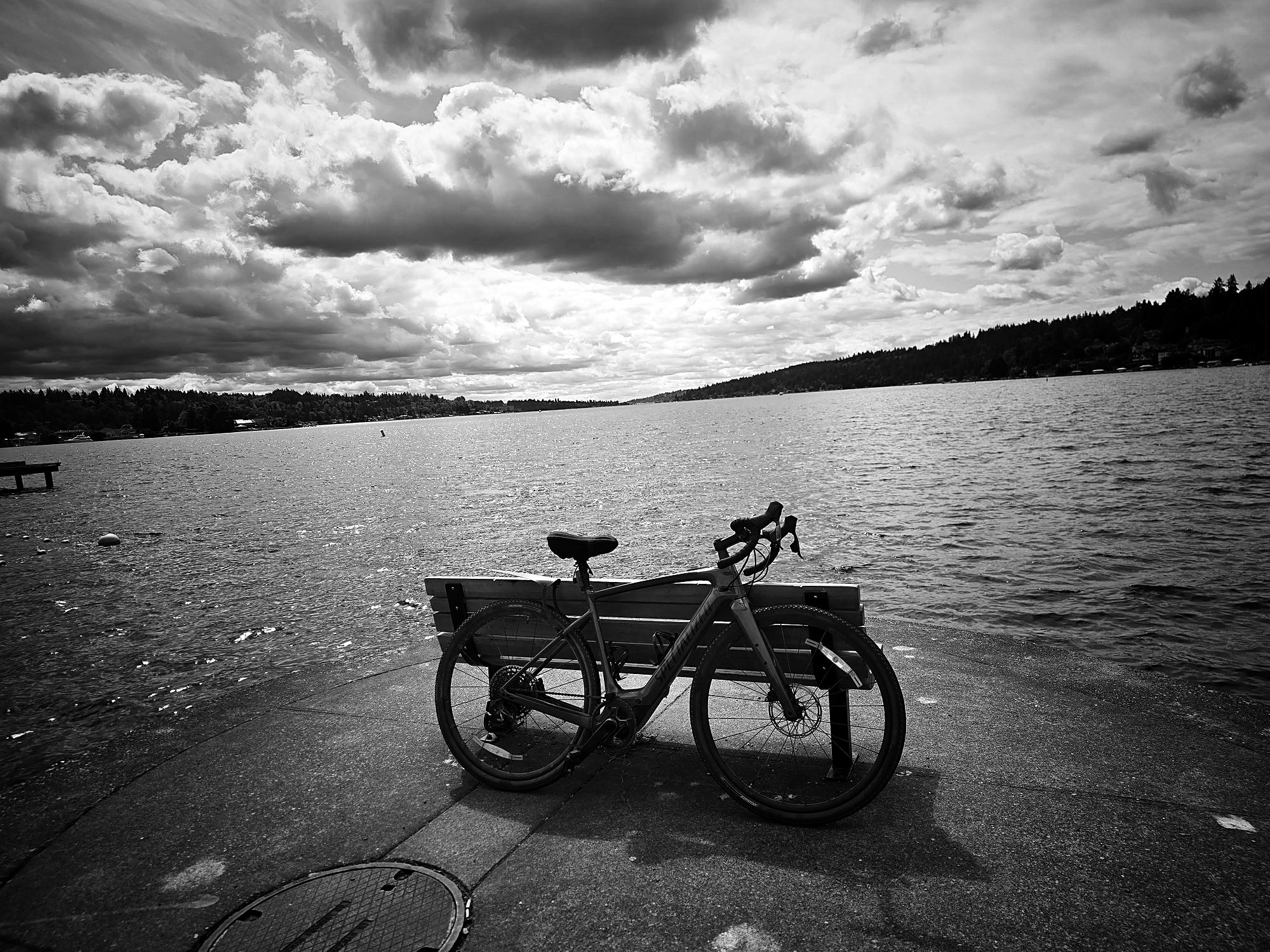 A gravel bike propped against a wooden bench at the edge of a lake under heavy cloud